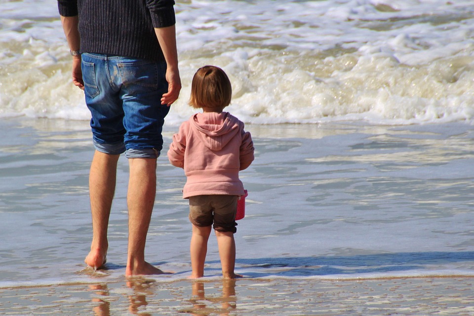 Erwachsener und Kleinkind stehen am Strand im flachen Wasser und schauen aufs Meer